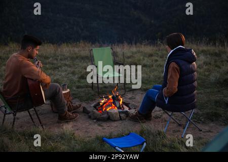 Coppia con chitarra seduta vicino falò nel campeggio Foto Stock