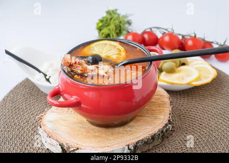 Solyanka di carne. Caldo combinato hodgepodge. una porzione di zuppa di carne rossa piccante con pomodori, salsicce affumicate e olive al limone in una turena rossa Foto Stock