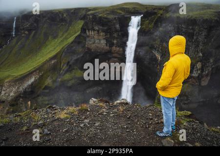 Un uomo in giacca gialla godendo la cascata Haifoss sulle piogge del tempo coperto. Highlands dell'Islanda. Foto Stock