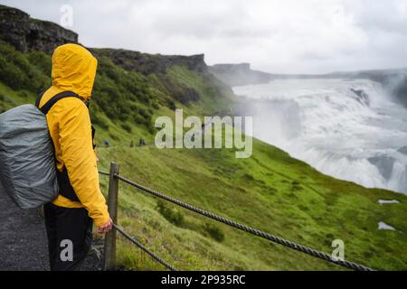 Turista con zaino e giacca verde visita Gullfoss potente famosa cascata in Islanda. Luogo famoso da visitare sulla rotta del cerchio d'Oro dell'Islanda. Foto Stock