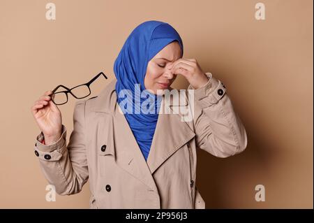 Donna musulmana araba multitasking iperlavorata in hijab blu e beige casual coat che intona occhiali, massaggiante fronte Foto Stock