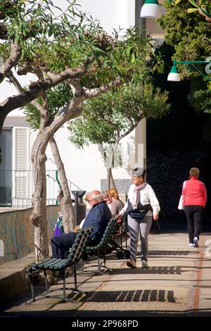 Persone che si godono il sole a piedi e sulle panchine, Capri. Capri è un'isola situata nel Mar Tirreno al largo della Penisola Sorrentina, sul lato sud del Golfo di Napoli, nel Campaniaregion d'Italia Capri, è famosa per il suo paesaggio aspro, la storia culturale, gli alberghi di lusso e lo shopping Foto Stock