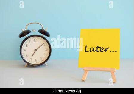 Concetto di procrastinazione, ritardo e urgenza. Sveglia bianca con una nota adesiva con testo in seguito Foto Stock