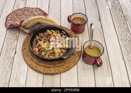 Uno stufato di manzo messicano cotto in una pentola di ghisa su tre quarti di sughero, tortilla di grano e salse Foto Stock