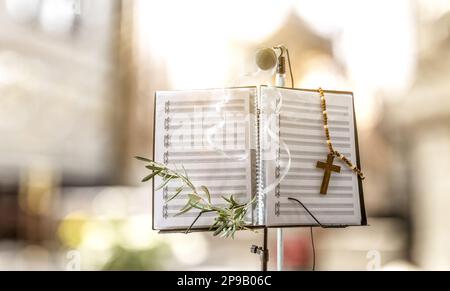 Concetto di canti corali per la celebrazione della Domenica delle Palme con stand musicale con spartito decorato con rametto di ulivo e croce religiosa e sfondo della chiesa. Foto Stock