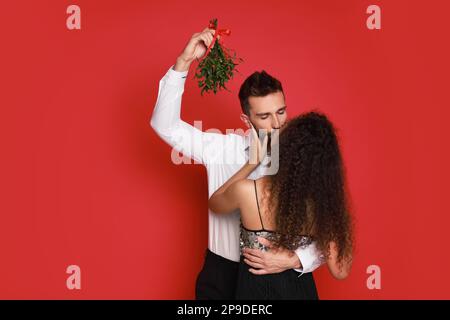 Felice coppia baciare sotto mazzo di mistletoe su sfondo rosso Foto Stock
