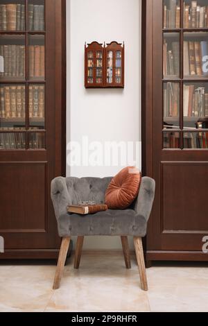 Comoda poltrona con cuscino e libro tra librerie in legno in biblioteca Foto Stock