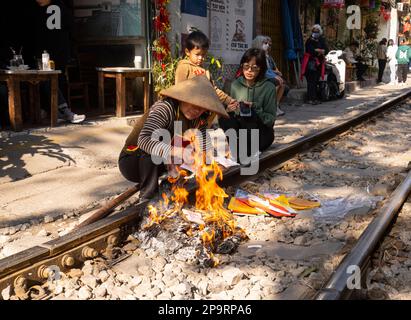 Hanoi, Vietnam. Jnauary 2023. Una donna che brucia tiền âm phủ, offerte votive per gli antenati lungo i binari del treno di fronte alla sua casa nel vecchio quarto Foto Stock