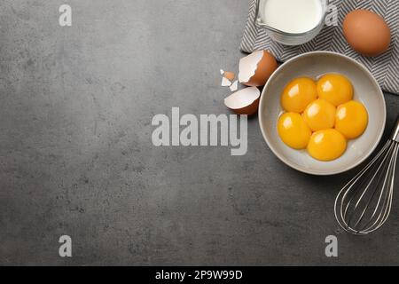 Ciotola con tuorli d'uovo crudi, frusta e caraffa di latte sul tavolo grigio, piatto. Spazio per il testo Foto Stock