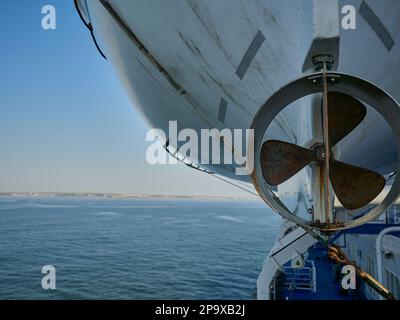 dettagli di un'elica di una scialuppa di salvataggio appesa sul lato di una barca che guida sul mare aperto in una chiara giornata di sole con cielo blu Foto Stock