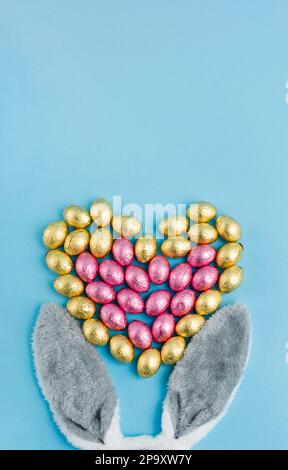 Orecchie di coniglio di Pasqua e mucchio di uova caramelle a forma di cuore avvolto in foglio rosa e dorato su sfondo blu. Buona Pasqua, amore concetto. Preparazione per Foto Stock