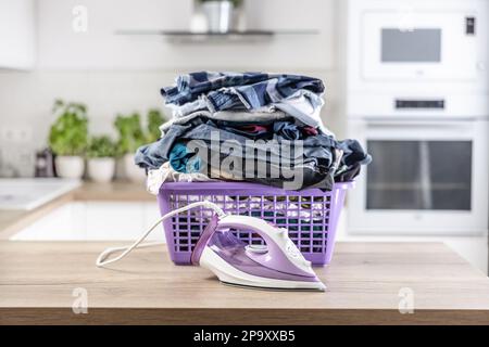 Un grande mucchio di vestiti stropicciati spilla un cestino viola su una scrivania da cucina con un ferro da stiro di fronte. Foto Stock