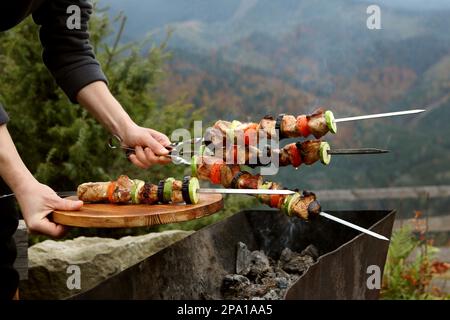 Donna che tiene spiedini di metallo e tavola con deliziosa carne contro il paesaggio di montagna, primo piano Foto Stock