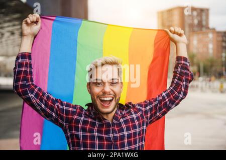 Felice uomo gay che indossa il trucco tenendo lgbt arcobaleno bandiera all'aperto - Focus on face Foto Stock