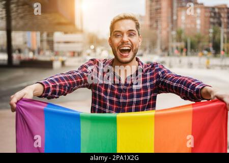 Felice uomo gay che indossa il trucco tenendo lgbt arcobaleno bandiera all'aperto - Focus on face Foto Stock