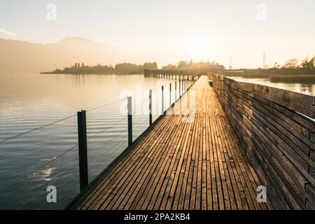 Una vista orizzontale di un lungo molo di legno sul mare in luce dorata sera con una silhouette di paesaggio montano sullo sfondo Foto Stock