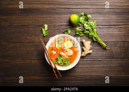 Zuppa tailandese tradizionale piccante e calda tom yum con gamberi, lime, coriandolo in una ciotola su sfondo rustico in legno, vista dall'alto. Sudore e acidore autentici Foto Stock