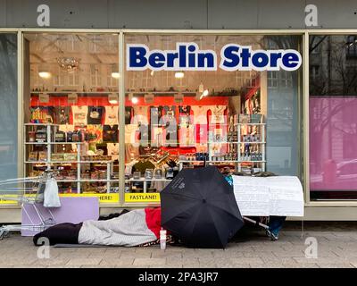 Senzatetto di fronte al 'Berlin Store', coperto da un ombrello dell'Hotel Adlon nel centro di Berlino, Germania nel febbraio 2023. Foto Stock