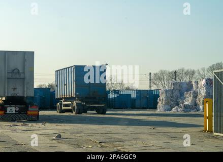 Rimorchio pesante parcheggiato nel cortile di riciclaggio lungo le balle di rifiuti in plastica e i contenitori in metallo blu. Foto Stock