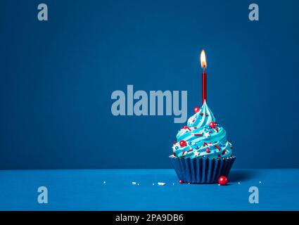 Cupcake di compleanno con crema di burro blu con spolverini rossi e candela su sfondo blu scuro. Foto Stock