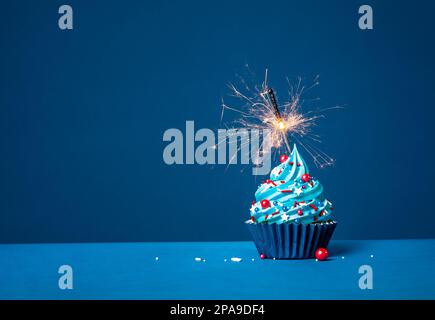 Cupcake festivo di compleanno con crema di burro rossa, bianca e blu e scintillante su sfondo blu scuro. Foto Stock