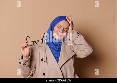 Elegante musulmana arabo bella donna in hijab blu, si tacca gli occhiali, le griche nel dolore, soffre di mal di testa tensione Foto Stock