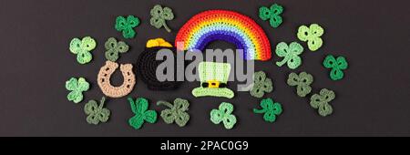 Concetto del giorno di San Patrizio. Composizione a maglia di un cappello verde, una pentola d'oro, un ferro di cavallo, un arcobaleno e shamrock verde su sfondo nero. Banner Foto Stock