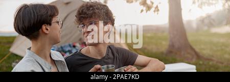 Giovane maschio e femmina seduto in natura vicino alla tenda, parlando e bevendo tè o caffè. Foto Stock