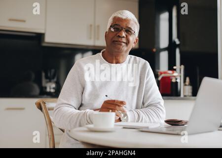Uomo d'affari maturo che guarda la fotocamera mentre lavora da casa. Uomo d'affari senior che utilizza un notebook e un computer portatile mentre lavora in remoto. Foto Stock