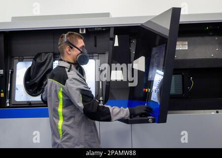 Stampante industriale a polvere 3D per metallo e operatore. Uomo in tuta protettiva vicino alla finestra di una grande stampante 3D in laboratorio. Le mani indossano grandi maniche nere per sigillare le lavorazioni all'interno della macchina Foto Stock