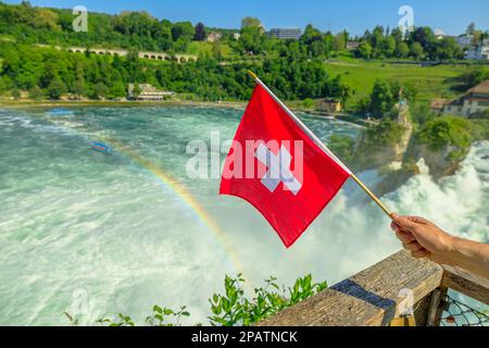 Primo piano di una bandiera svizzera con arcobaleno. Veduta aerea della cascata svizzera delle cascate del Reno in Svizzera. La cascata più grande e potente d'Europa Foto Stock
