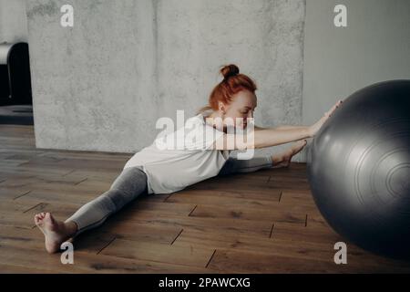 Allenamento stretching. Giovane donna in forma e sportiva che si esercita con il fitness in studio mentre si siede in posizione divisa a piedi nudi sul pavimento, pratica Foto Stock