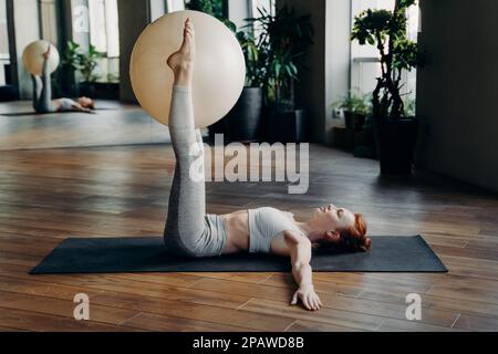 Giovane donna sportiva in abbigliamento sportivo grigio sdraiato sul tappetino yoga in palestra e facendo esercizi di stabilità palla, tenendo il fit tra le gambe A. Foto Stock