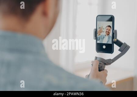 Vista posteriore del bel giovane uomo brunete sorridente con stoppia facendo selfie su smartphone con stabilizzatore gimbal a casa e gesturing segno ok, indossa c Foto Stock