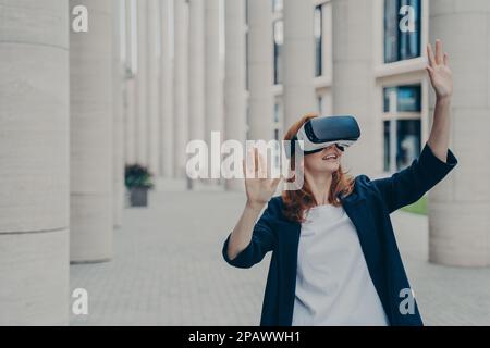 La realtà virtuale come strumento per la vostra azienda. Giovane donna zenzero vestita formalmente in piedi all'aperto e utilizzando visore VR o occhiali di protezione, interagendo con int Foto Stock