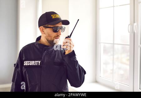 Guardia di sicurezza in uniforme in piedi all'interno della casa, guardando fuori dalla finestra, e parlando alla radio Foto Stock