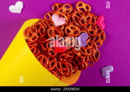 Mini pretzel e piccoli cuori colorati in cono di carta giallo giallano su sfondo viola primo piano. Concetto di pretzel di amore Foto Stock