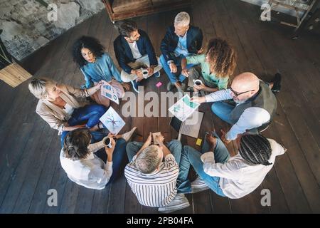 Un gruppo diversificato di professionisti si impegna in una sessione di brainstorming creativo, seduti sul pavimento e condividendo idee. Con notebook, tablet e notebook Foto Stock