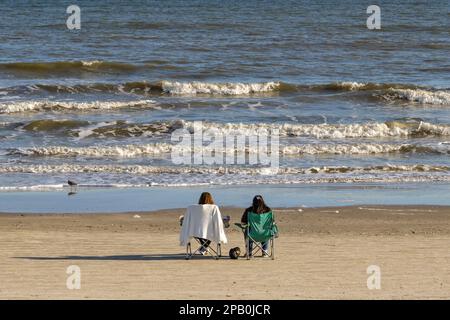 Galveston, Texas, USA - Febbraio 2023: Due persone che si rilassano sulle sedie sul bordo dell'acqua guardando verso il mare Foto Stock