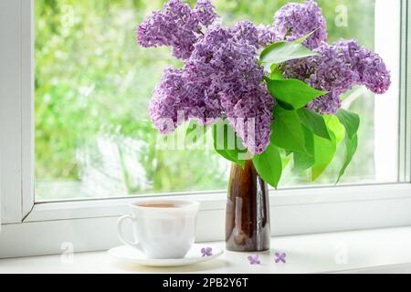 Interno domestico con un bouquet di fiori di lilla fiorenti sulla finestra. Festa del tè in primavera mattina in cucina vicino alla finestra Foto Stock