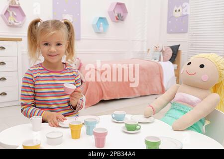 Simpatica bambina che gioca a una festa con bambola al tavolo in camera, spazio per il testo Foto Stock