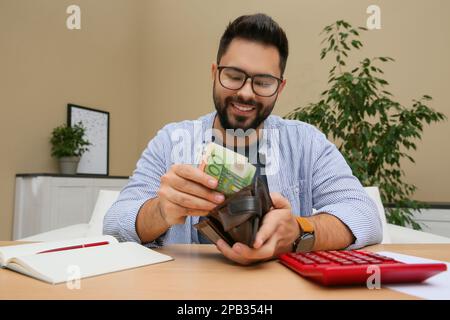 Giovane uomo che mette i soldi nel portafoglio al tavolo all'interno Foto Stock