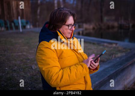 Una donna legge i messaggi sul suo smartphnoe mentre si trova accanto ad un Lake.Late nel pomeriggio. Foto Stock