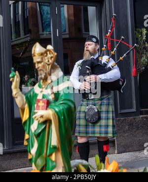 Warrington, Cheshire, Regno Unito - 12 marzo 2023 – il Warrington Irish Club ospita il St. La parata del giorno di Patrick. A partire dalle 10,30 del mattino dall'Irish Club di Orford Lane fino al "River of Life" di Bridge Street, nel centro della città, dove si è svolto un breve servizio per ricordare l'anniversario del bombardamento di Warrington. Un solitario suonò vicino a una statua di San Patrizio Credit: John Hopkins/Alamy Live News Foto Stock
