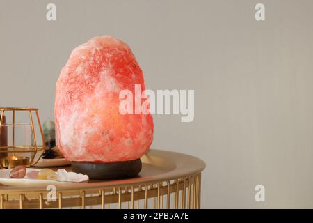 Lampada di sale Himalayan, candela e cristalli su tavolo dorato vicino a parete grigio chiaro, spazio per il testo Foto Stock