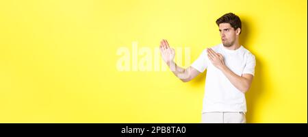 Uomo che mostra abilità kung-fu, arti marziali movimento ninja, in piedi in bianco t-shirt pronto a combattere, in piedi su sfondo giallo Foto Stock