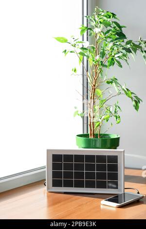 Pannello solare portatile che carica telefono cellulare e impianto domestico sul tavolo vicino alla finestra Foto Stock