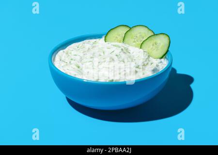 Salsa tzatziki fatta in casa in una ciotola blu in luce brillante su un tavolo dai colori vivaci. Ciotola con salsa tzatziki isolata su fondo blu. Foto Stock