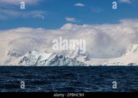 Vista sull'oceano della costa della penisola antartica. Montagne con neve e ghiaccio, sotto uno strato spesso di nuvole. Oceano blu scuro in primo piano; cielo blu A. Foto Stock