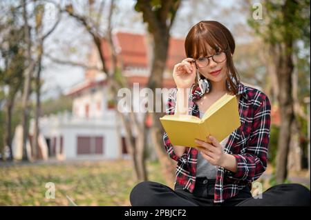 Bella giovane studentessa asiatica in abiti casual sta toccando i suoi occhiali mentre si concentra sulla lettura di un libro su una panchina nel parco del campus. Foto Stock